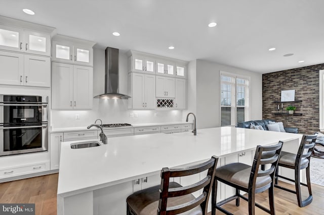 kitchen with light wood finished floors, wall chimney exhaust hood, a center island with sink, and appliances with stainless steel finishes