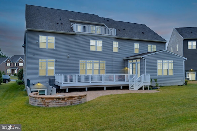 back of house at dusk with a balcony, a wooden deck, a yard, and roof with shingles