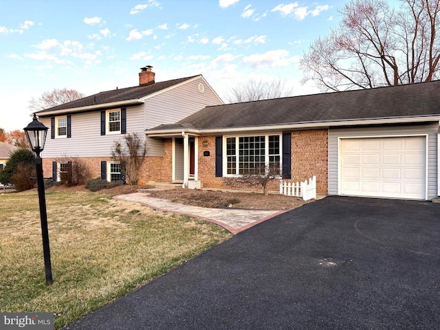 split level home featuring aphalt driveway, a front yard, an attached garage, brick siding, and a chimney