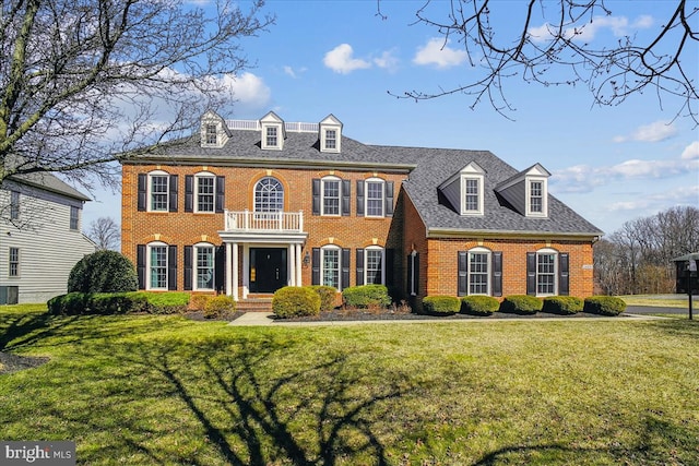 georgian-style home featuring a front yard, a balcony, brick siding, and a shingled roof