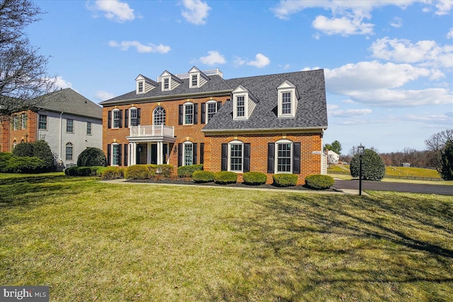 colonial house featuring a front yard, a balcony, roof with shingles, and brick siding