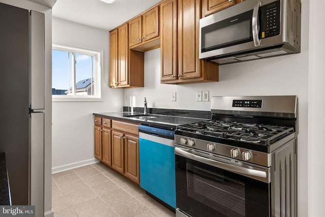 kitchen with a sink, dark countertops, appliances with stainless steel finishes, brown cabinetry, and baseboards