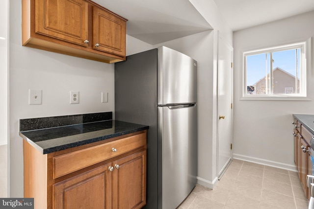 kitchen featuring freestanding refrigerator, dark stone counters, brown cabinetry, light tile patterned floors, and baseboards