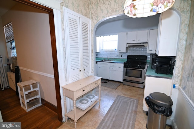 kitchen featuring stainless steel gas range oven, wallpapered walls, baseboards, under cabinet range hood, and white cabinets