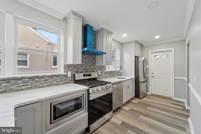 kitchen with light wood-style flooring, ornamental molding, a sink, appliances with stainless steel finishes, and wall chimney exhaust hood