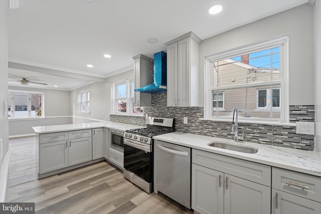 kitchen featuring light wood finished floors, a peninsula, stainless steel appliances, wall chimney exhaust hood, and a sink