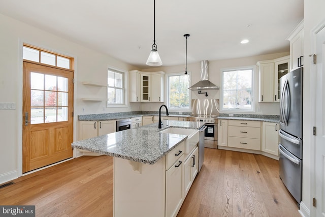 kitchen with glass insert cabinets, wall chimney exhaust hood, light wood-style floors, and stainless steel appliances