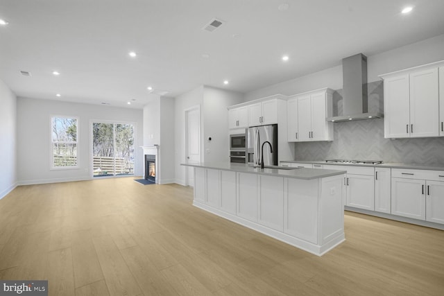 kitchen with visible vents, a kitchen island with sink, stainless steel appliances, white cabinetry, and wall chimney range hood