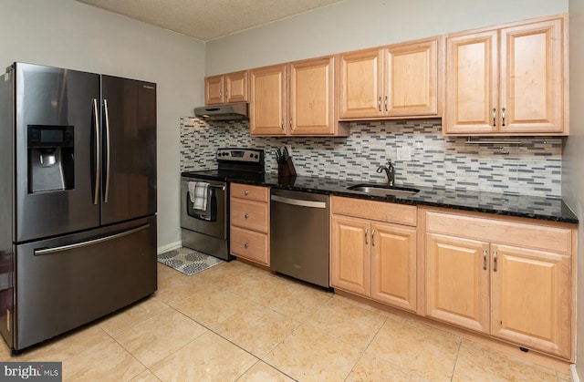 kitchen with light brown cabinetry, a sink, under cabinet range hood, dark stone counters, and appliances with stainless steel finishes