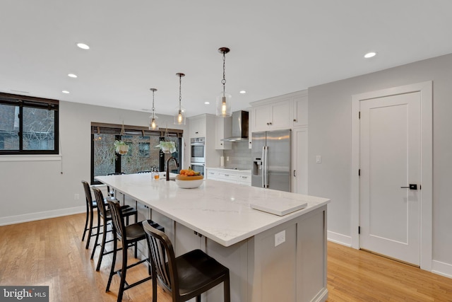 kitchen featuring wall chimney range hood, light wood-type flooring, a large island with sink, and stainless steel appliances