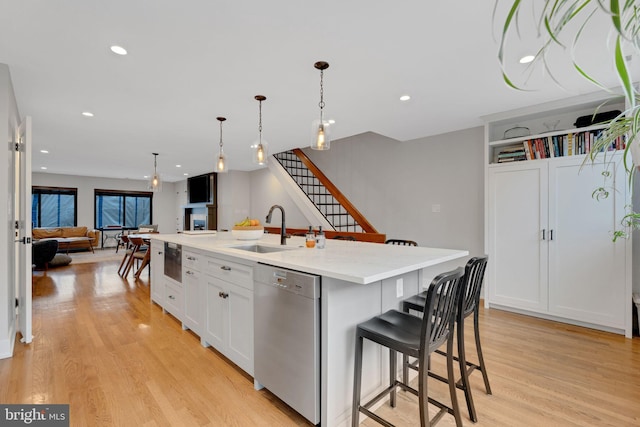 kitchen featuring light wood-style flooring, a kitchen island with sink, a sink, stainless steel dishwasher, and open floor plan