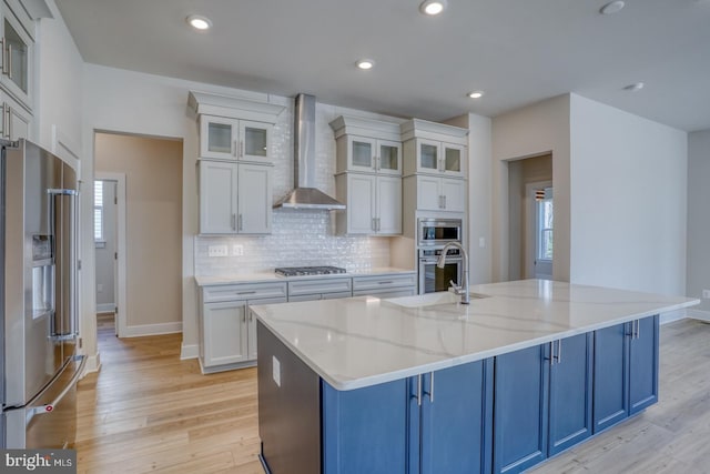 kitchen with blue cabinetry, light wood-style flooring, stainless steel appliances, wall chimney exhaust hood, and backsplash