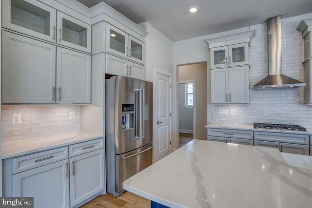 kitchen featuring wall chimney range hood, stainless steel appliances, light wood finished floors, decorative backsplash, and light stone countertops