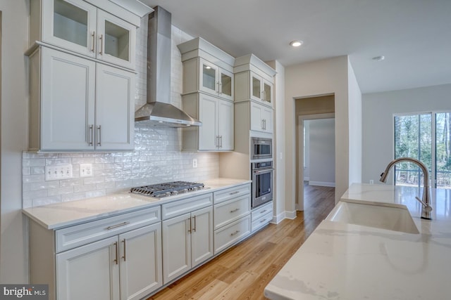 kitchen featuring light stone counters, a sink, appliances with stainless steel finishes, wall chimney range hood, and backsplash