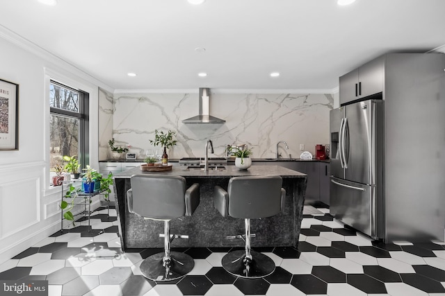 kitchen featuring a center island with sink, stainless steel refrigerator with ice dispenser, wall chimney range hood, crown molding, and dark floors