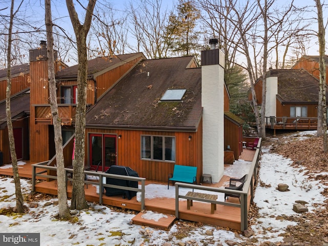 snow covered house with a balcony, a deck, a chimney, and a shingled roof