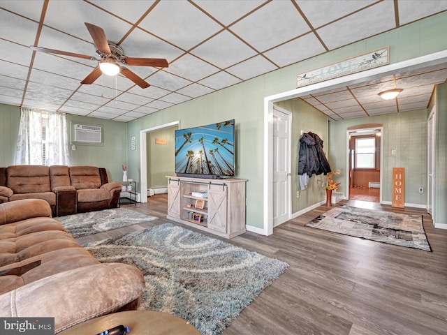 living room with a wall unit AC, wood finished floors, baseboards, ceiling fan, and a paneled ceiling