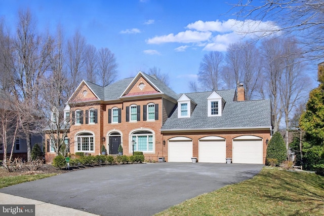 colonial inspired home featuring driveway, an attached garage, a shingled roof, brick siding, and a chimney