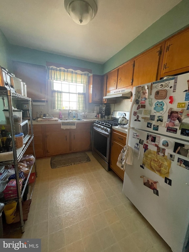 kitchen featuring stainless steel gas stove, under cabinet range hood, a sink, freestanding refrigerator, and brown cabinetry