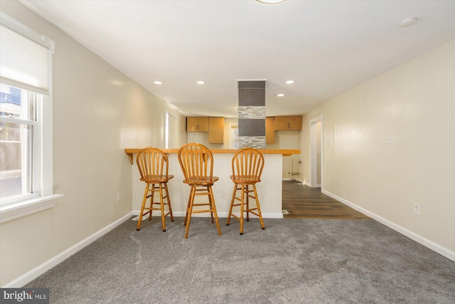 kitchen featuring baseboards, a peninsula, recessed lighting, a kitchen bar, and dark colored carpet