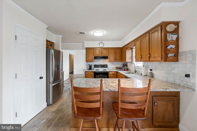 kitchen featuring under cabinet range hood, electric range oven, brown cabinets, a peninsula, and freestanding refrigerator