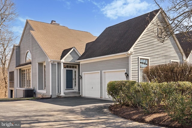 cape cod-style house with an attached garage, central AC, a chimney, a shingled roof, and aphalt driveway