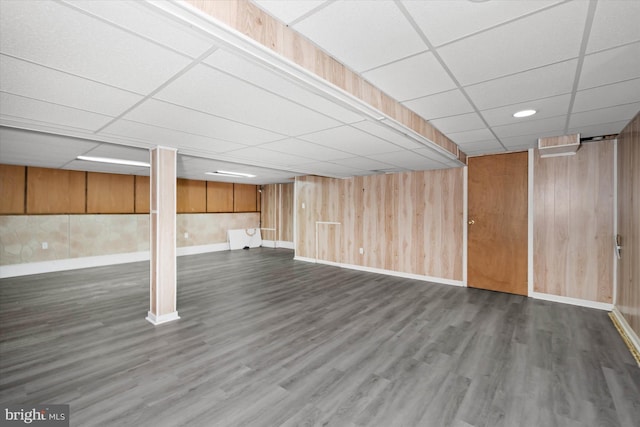 finished basement featuring a drop ceiling, baseboards, wood walls, and dark wood-style flooring