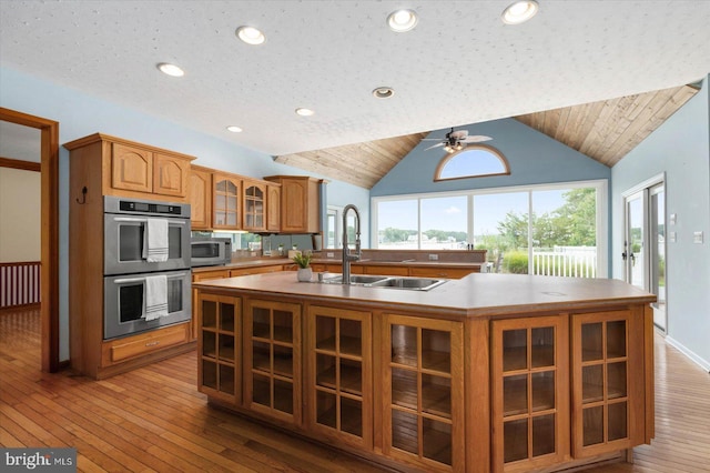 kitchen featuring glass insert cabinets, ceiling fan, an island with sink, brown cabinets, and appliances with stainless steel finishes