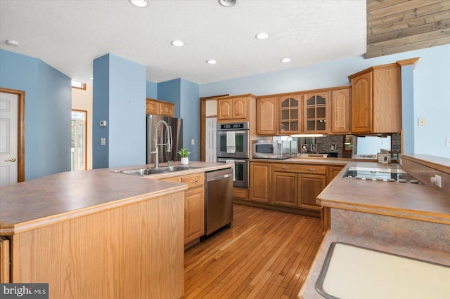 kitchen featuring an island with sink, a sink, appliances with stainless steel finishes, light wood finished floors, and glass insert cabinets