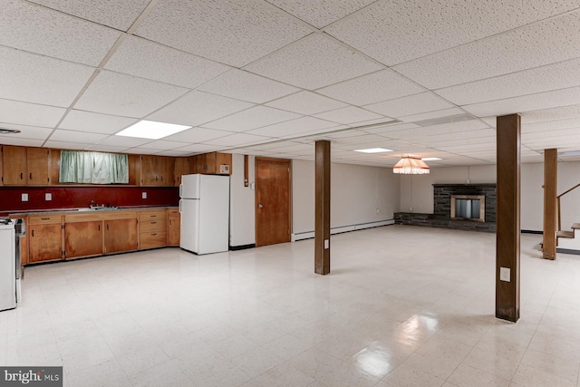 finished basement with a drop ceiling, light floors, freestanding refrigerator, a stone fireplace, and baseboard heating