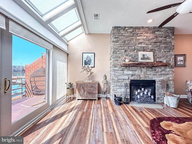 interior space with a ceiling fan, wood finished floors, vaulted ceiling with skylight, a stone fireplace, and baseboards