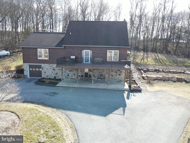 view of front of house featuring aphalt driveway, a wooden deck, roof with shingles, a patio area, and an attached garage