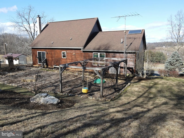 rear view of property featuring solar panels, a yard, a shingled roof, a chimney, and a deck