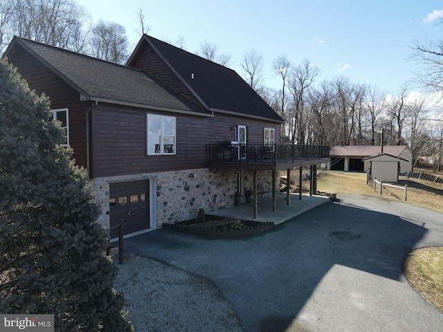 view of side of property featuring roof with shingles, a garage, a deck, stone siding, and driveway