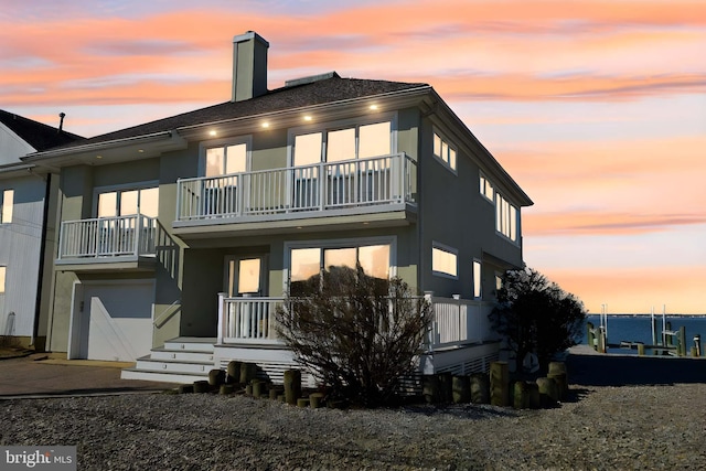 back of house with concrete driveway, stucco siding, a chimney, a balcony, and an attached garage