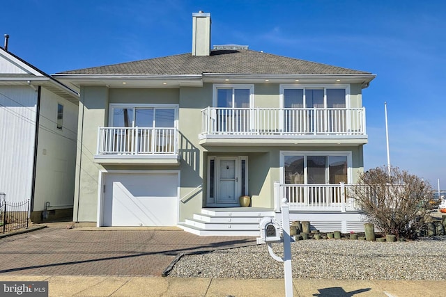 view of front of home with a balcony, an attached garage, a chimney, stucco siding, and decorative driveway