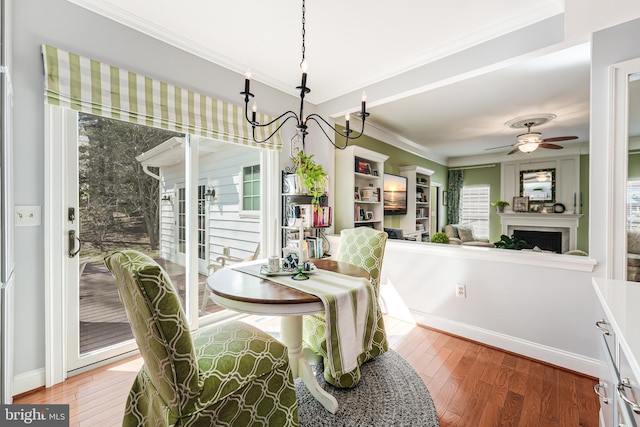 dining room featuring crown molding, baseboards, hardwood / wood-style floors, ceiling fan with notable chandelier, and a fireplace