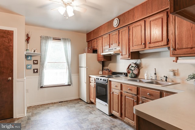 kitchen featuring under cabinet range hood, wainscoting, brown cabinets, white appliances, and a sink