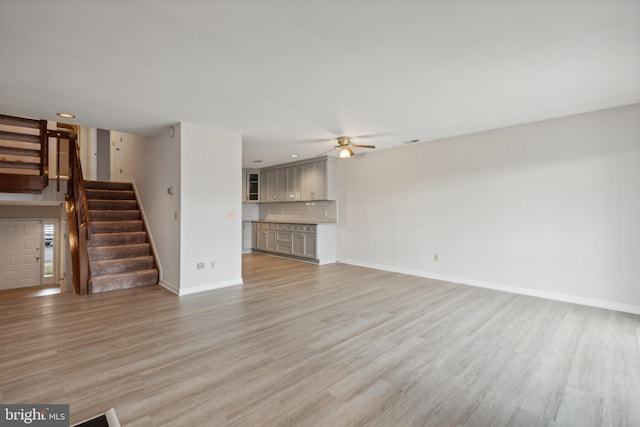 unfurnished living room featuring stairs, baseboards, light wood finished floors, and ceiling fan