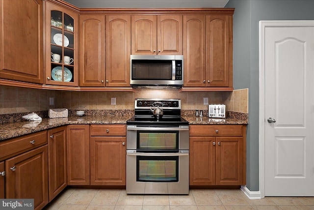 kitchen featuring backsplash, glass insert cabinets, dark stone countertops, appliances with stainless steel finishes, and brown cabinets