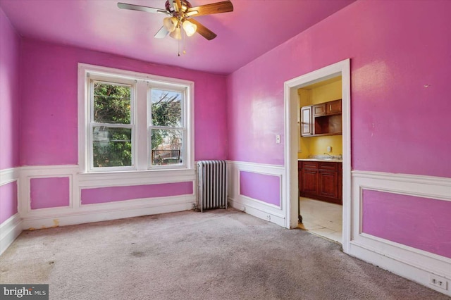 carpeted empty room featuring a sink, a wainscoted wall, a ceiling fan, and radiator heating unit