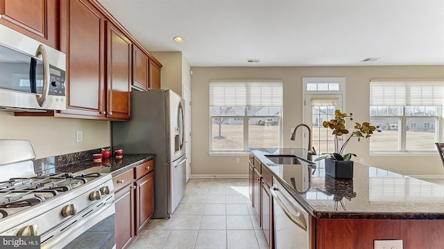 kitchen featuring a sink, dark stone counters, light tile patterned floors, appliances with stainless steel finishes, and a kitchen island with sink