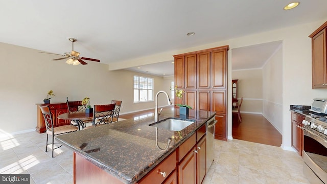 kitchen with a sink, ceiling fan, dark stone counters, stainless steel appliances, and a kitchen island with sink