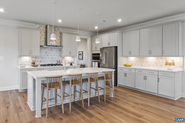 kitchen with wall chimney range hood, light countertops, light wood-style flooring, a kitchen breakfast bar, and appliances with stainless steel finishes