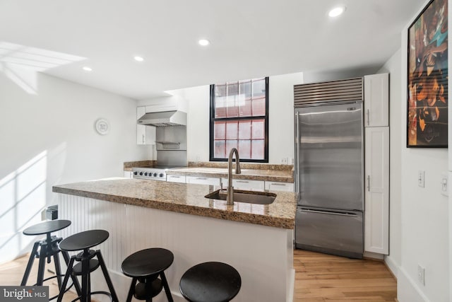 kitchen featuring light wood finished floors, stainless steel built in refrigerator, a kitchen breakfast bar, wall chimney exhaust hood, and a sink