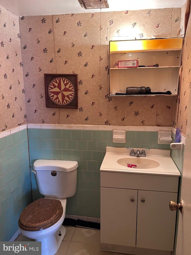 half bath with wallpapered walls, a wainscoted wall, toilet, tile patterned floors, and vanity