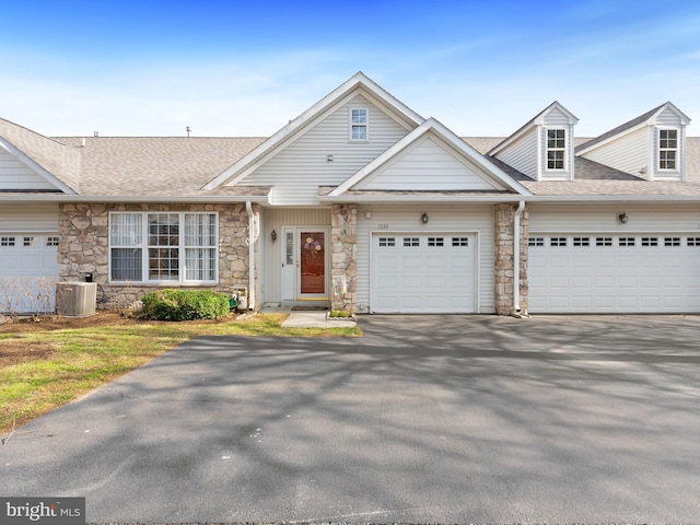 townhome / multi-family property featuring roof with shingles, a garage, stone siding, aphalt driveway, and central air condition unit