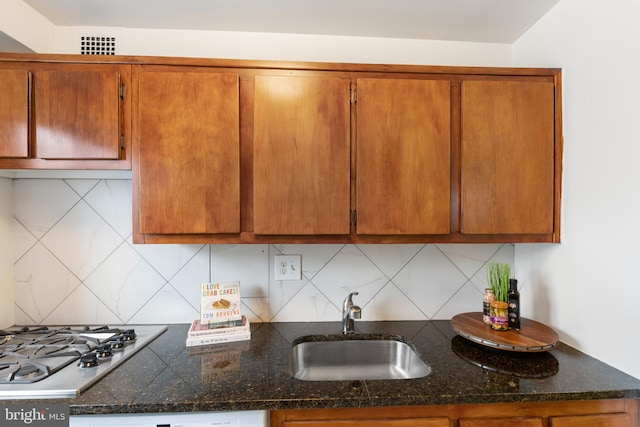 kitchen with tasteful backsplash, dark stone counters, brown cabinets, stainless steel gas stovetop, and a sink