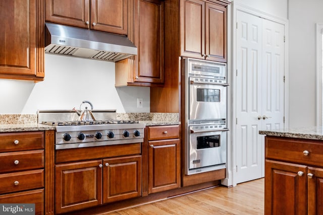 kitchen featuring brown cabinets, under cabinet range hood, light stone counters, stainless steel appliances, and light wood finished floors