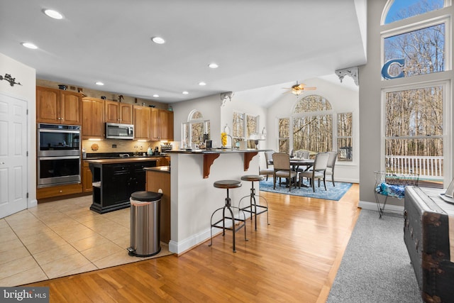 kitchen featuring a ceiling fan, a center island with sink, appliances with stainless steel finishes, dark countertops, and a kitchen breakfast bar